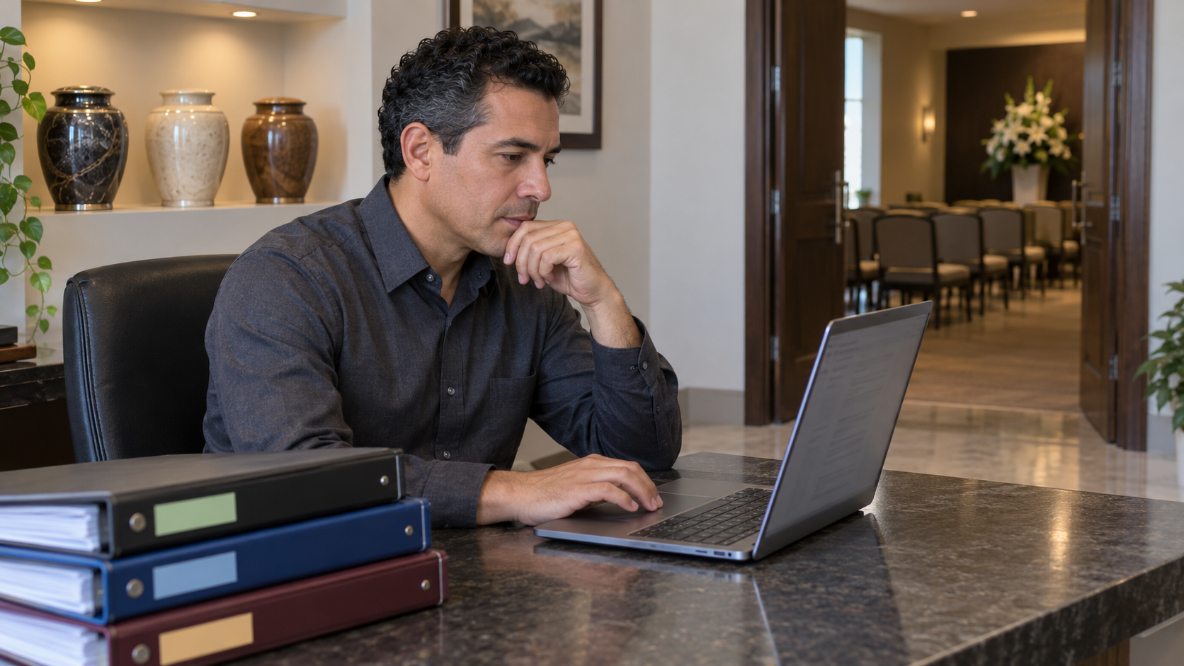 Hombre revisa fuentes en una laptop dentro de un área administrativa de funeraria con urnas de muestra y un salón vacío al fondo.